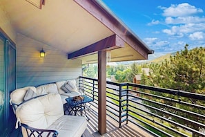 Ein überdachter Balkon mit einer Couch und einem Tisch, einem Geländer und Blick auf eine hügelige Landschaft.