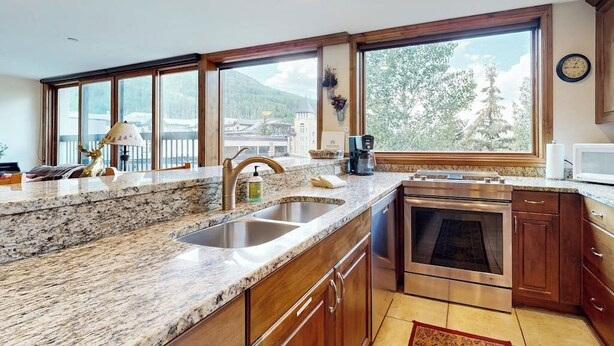 A kitchen with granite countertops, stainless steel appliances, wooden cabinets, and large windows offering a view of a mountain and a building. - Vail Spa Condo w/ Pools, Hot Tubs & Gorgeous Valley Views - Near Lifts (Vail)