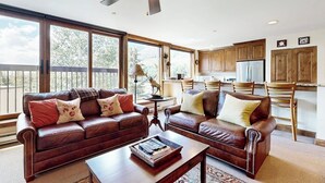 A spacious living room with a large window, a ceiling fan, and a kitchen area with wooden cabinets. - Vail Spa Condo w/ Pools, Hot Tubs & Gorgeous Valley Views - Near Lifts (Vail)