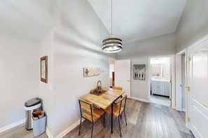 Spiseplads med et træbord, stole, en hængende lampe og en skraldespand.