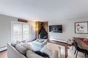 A living room with a fireplace, a sofa, a dining table, and a TV mounted on the wall. - Mountain Views at Vail East | Fireplace, Desk, Walk to Vail Shuttle & Trails (Vail)