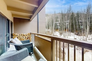 A balcony with wicker furniture, a stack of firewood, and a view of a snowy landscape with trees and buildings. - Ideally located condo with pool & hot tub, next to Vail Golf Club/Nordic Center (Vail)