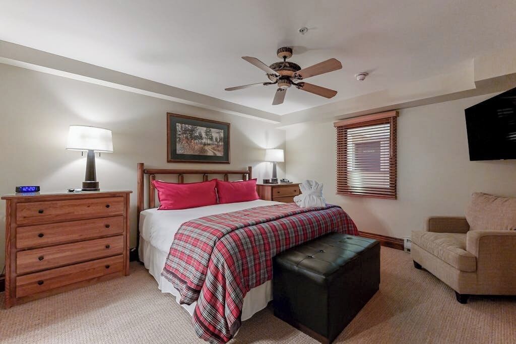 A bedroom with a bed, bedside tables, a nightstand, a ceiling fan, a window with blinds, a TV, and a sofa.