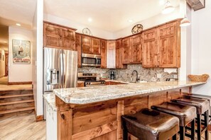 A kitchen with wooden cabinets, stainless steel appliances, a marble countertop, and bar stools. - Ground Floor Mountain Condo with Shared Pool & Hot Tub - Free Ski Shuttle (Avon)