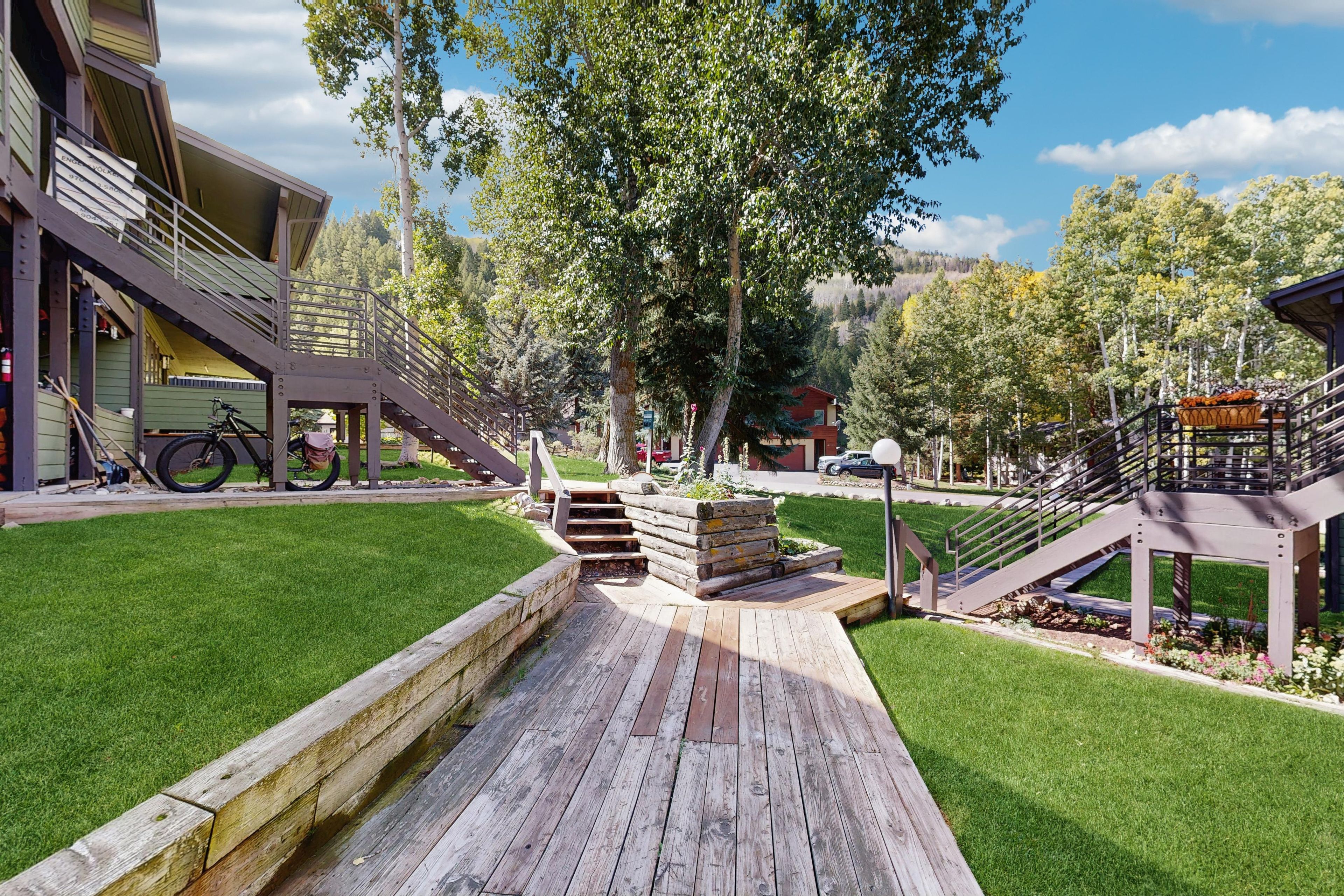 A wooden deck with a staircase leading to a building, surrounded by a well-maintained lawn and trees.