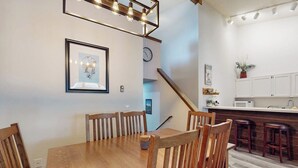 Een eetgedeelte met een houten tafel en stoelen, een keuken met witte kasten, en een trap naar een ander niveau.