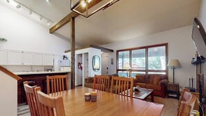 Een moderne keuken met witte kastjes, een eethoek met een houten tafel en stoelen, en een woonkamer met een bank en een groot raam.