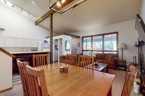 Eine moderne Küche mit weißen Schränken, ein Essbereich mit einem Holztisch und Stühlen sowie ein Wohnzimmer mit einer Couch und einem großen Fenster.