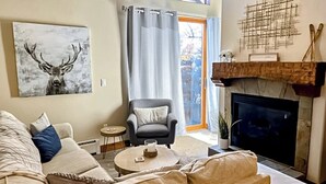 Una sala de estar con chimenea, un sofá, un sillón, un cuadro de un ciervo y una ventana con cortinas.