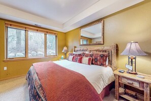 Ein Schlafzimmer mit einem großen Bett, einem Nachttisch mit Lampe, einem Spiegel über dem Bett und einem Fenster mit Blick auf Bäume.