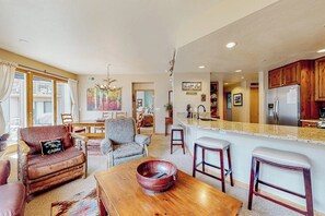Ein geräumiges Wohnzimmer mit Couch, Sessel und Couchtisch. Es gibt eine Küchenzeile mit Holzschränken und einem Kühlschrank.