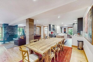 A spacious living area with a wooden dining table, a stone fireplace, and a kitchen in the background. - Charming Vail Condo with Fireplace, Balcony Views, Pool & Hot Tub (Vail)
