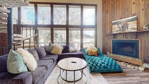 Un salon moderne avec un canapé d’angle, une table basse ronde, une cheminée et une grande fenêtre donnant sur la forêt.