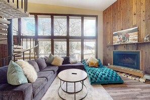 Ein modernes Wohnzimmer mit einer Eckcouch, einem runden Couchtisch, einem Kamin und einem großen Fenster mit Blick auf Bäume.