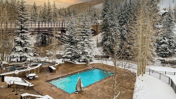 Un área de piscina con cubierta de madera, rodeada de árboles nevados y montañas al fondo.