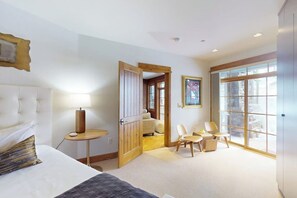 Ein Hotelzimmer mit einem Bett, einem Nachttisch mit Lampe, einer Holztür, einem kleinen Tisch mit Stühlen und großen Fenstern.
