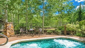 A hot tub surrounded by a stone wall, with a wooden bench and chairs, and a metal fence enclosing the area. - Ski In ski out condo with fully stocked kitchen, wifi, and shared hot tub (Avon)