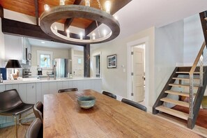 A dining area with a wooden table, chairs, and a large bowl. There is a kitchen in the background with cabinets and appliances. A staircase is visible to the right. - Colorful Riverfront Chalet  with Shared Pool and Hot Tub - Close to Ski Slopes (Vail)
