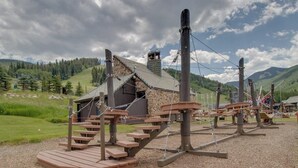 Eine rustikale Holzhütte mit einem Steinhaus im Hintergrund, umgeben von einer Wiese und Bergen.