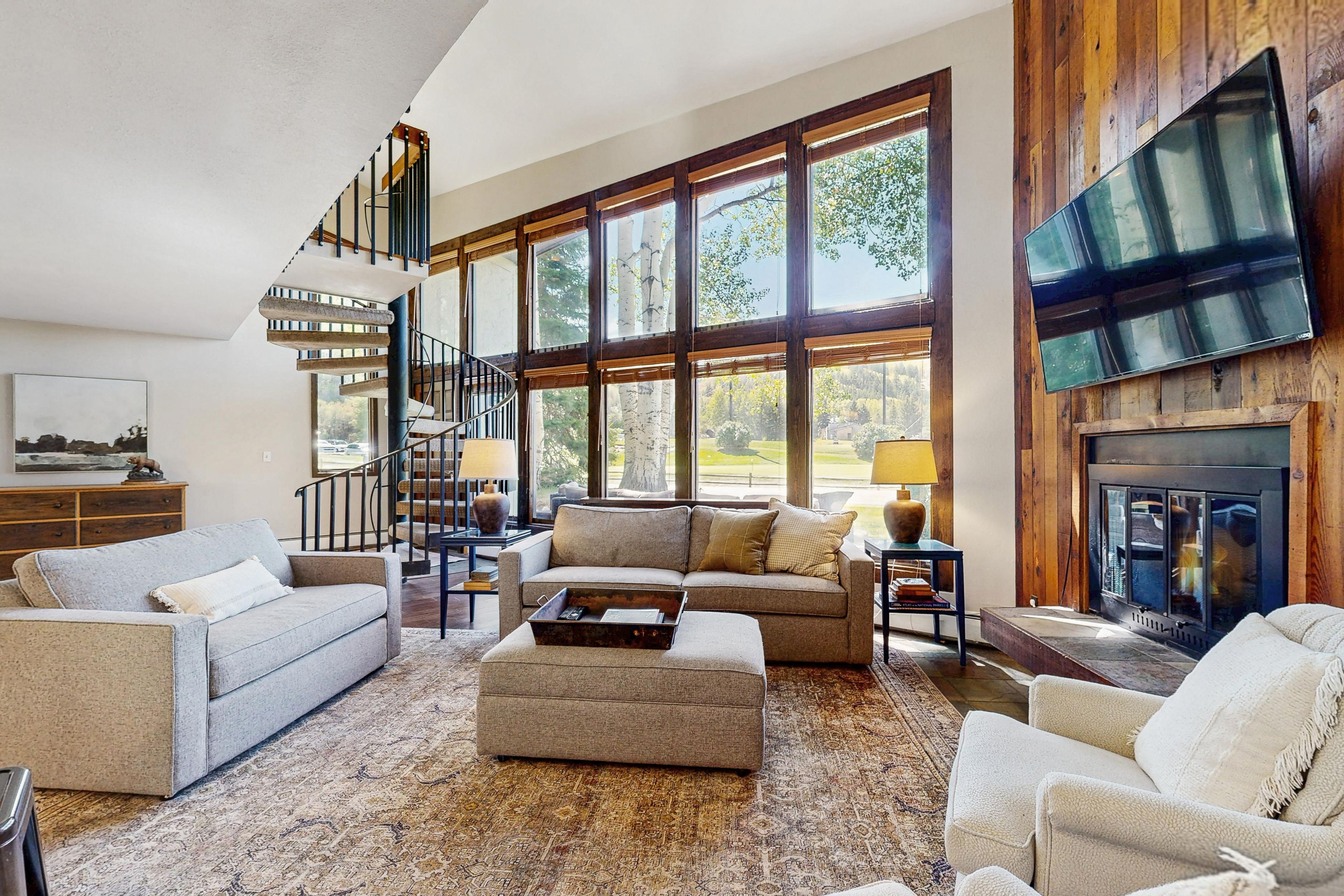 A spacious living room with a large window, a fireplace, a staircase, and a flat-screen TV mounted on the wall.