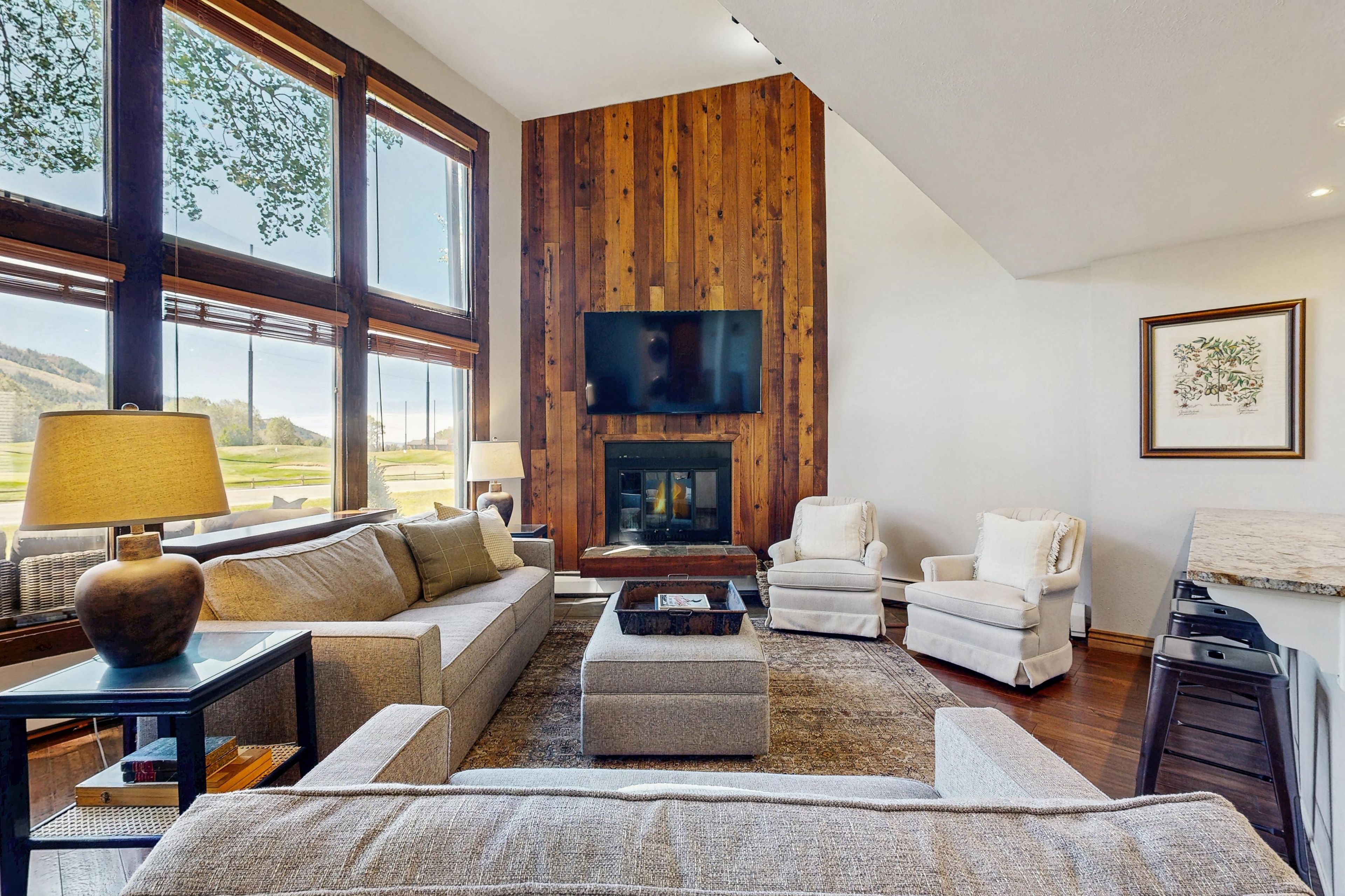 A spacious living room with a fireplace, large windows, and a high wooden wall.