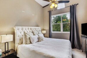Ein Schlafzimmer mit einem gesteppten Kopfteil, einem Bett mit weißer Bettdecke, einem Nachttisch mit Lampe, einem Fenster mit Vorhängen und einem Deckenventilator.