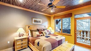 A bedroom with a wooden headboard, a quilted bedspread, a nightstand with a lamp, a bench, and a window with a view of trees.