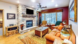 A living room with a stone fireplace, a ceiling fan, and a large window.
