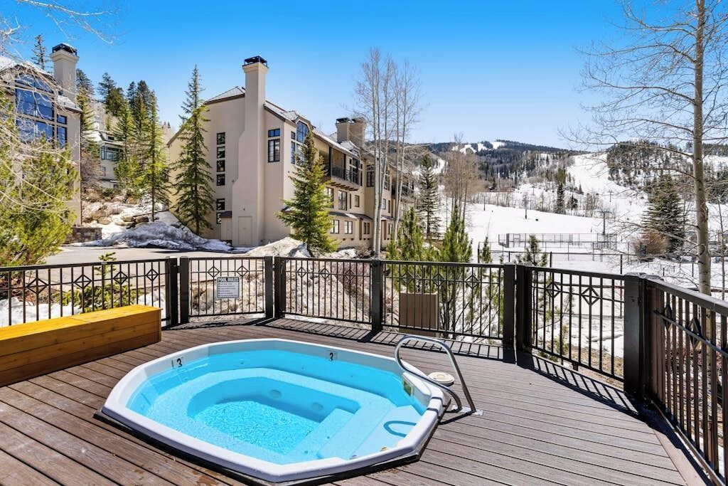 A wooden deck with a hot tub, a view of a snowy landscape, and a building with multiple windows and chimneys.