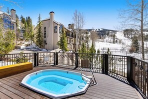 Eine Holzterrasse mit Whirlpool, Blick auf eine verschneite Landschaft und ein Gebäude mit zahlreichen Fenstern und Schornsteinen.
