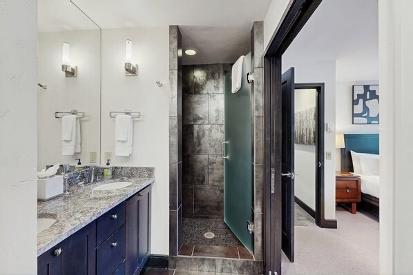 A modern bathroom with a marble countertop, a shower, and a door leading to a bedroom.