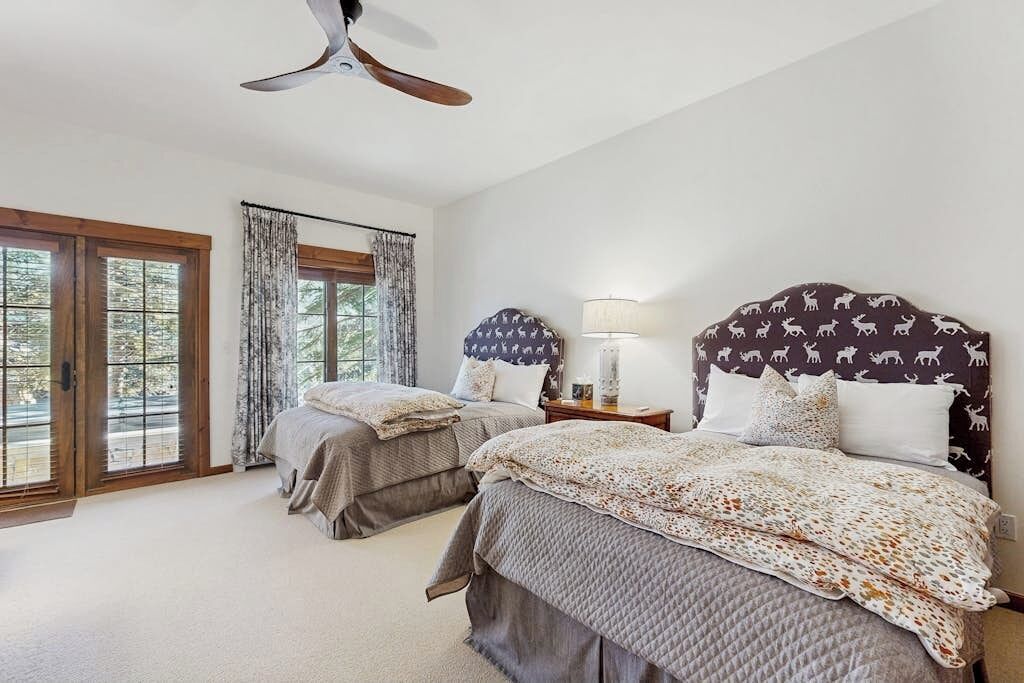 A bedroom with two beds, a ceiling fan, a window with curtains, and a lamp on a bedside table.