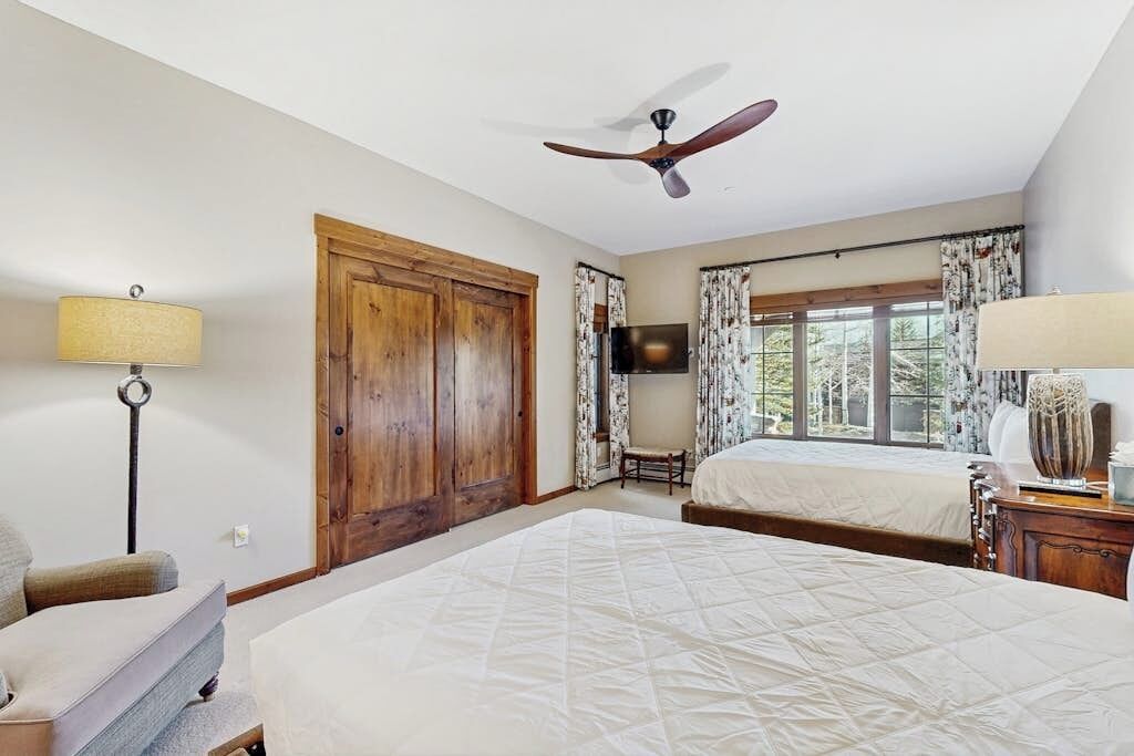 Quarto de hotel com duas camas, um ventilador de teto, uma televisão, uma janela com cortinas e um guarda-roupa de madeira.