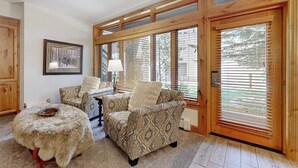 Ein gemütliches Wohnzimmer mit einem gemusterten Sofa, einem runden, mit Fell bezogenen Hocker und großen Fenstern mit Jalousien.