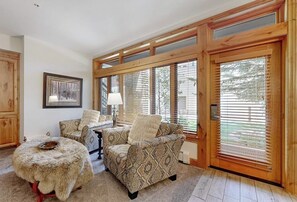 Ein gemütliches Wohnzimmer mit einem gemusterten Sofa, einem runden, mit Fell bezogenen Hocker und großen Fenstern mit Jalousien.