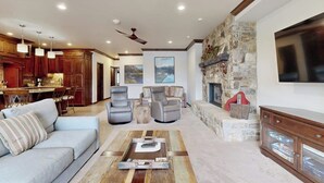 Amplia sala de estar con un sofá, dos sillones, una mesa de centro, chimenea, pared de piedra y una cocina al fondo.