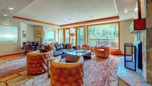 A spacious living room with a large sofa, ottoman, and coffee table, large windows offering a view of the outdoors, and a stone fireplace. - Ski-in/out Lionshead retreat w/ fireplace, fast WiFi, W/D & shared pool/hot tubs (Vail)