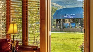 Vista attraverso una finestra con persiane di legno, una lampada e un tavolo con un oggetto decorativo. Si vede all'esterno un edificio con la scritta 'Eagle Bahn' e tetto blu, una zona erbosa e un paesaggio collinare sullo sfondo.