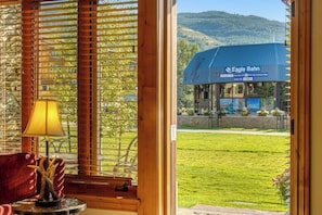 A view through a window with wooden blinds, a lamp, and a table with a decorative item. Outside, there's a building labeled 'Eagle Bahn' with a blue roof, a grassy area, and a hilly landscape in the background. - Ski-In/Ski-Out Villa w/ Fireplace, High-Speed WiFi & Shared Hot Tub, Pool, & W/D (Vail)