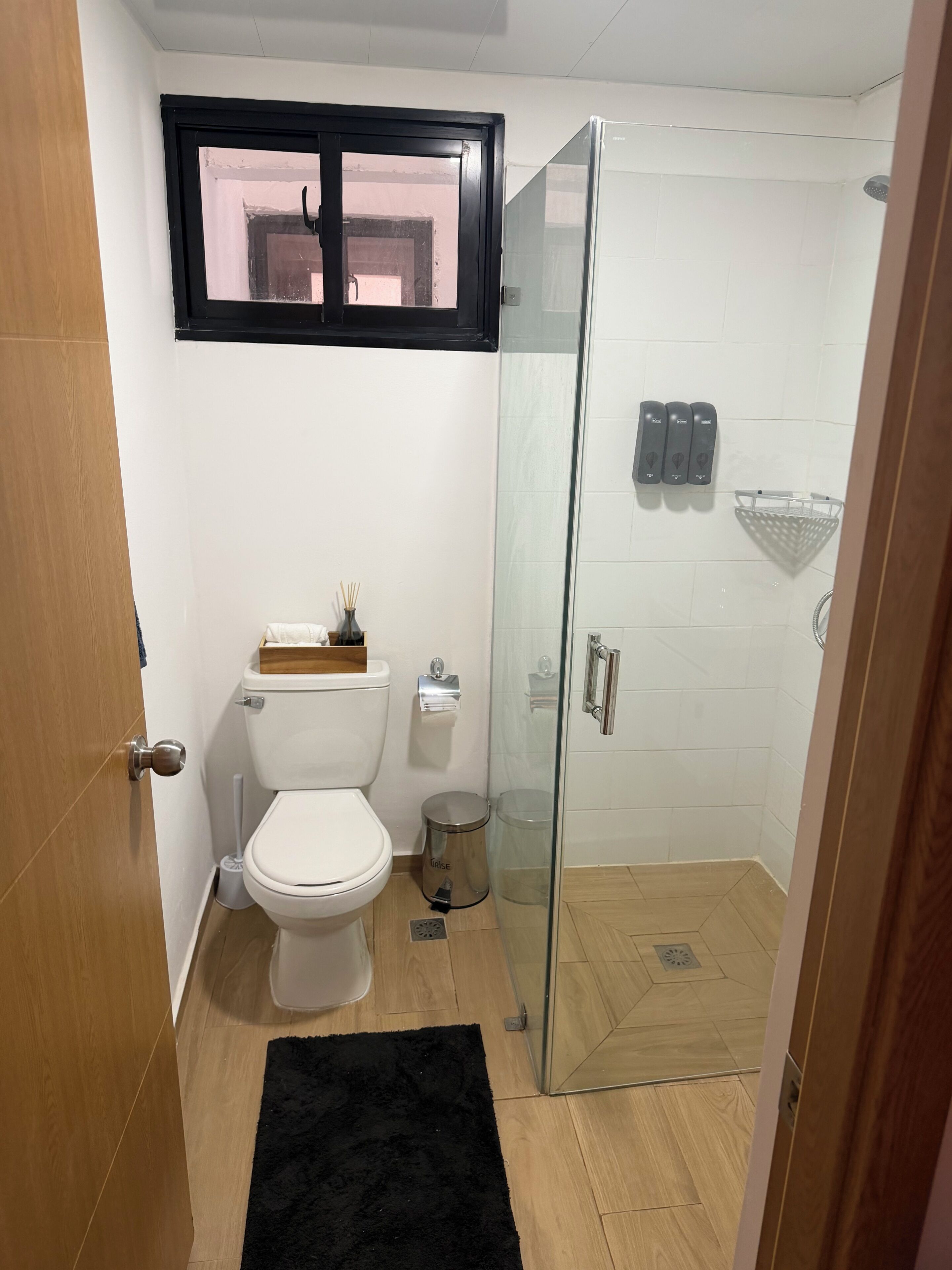Un baño con inodoro, cabina de ducha de vidrio, alfombra negra y ventana sobre el inodoro.