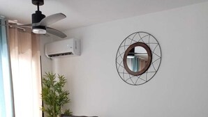 Una sala de estar con un sofá gris, dos otomanes tejidos, un espejo redondo y una planta de bambú.