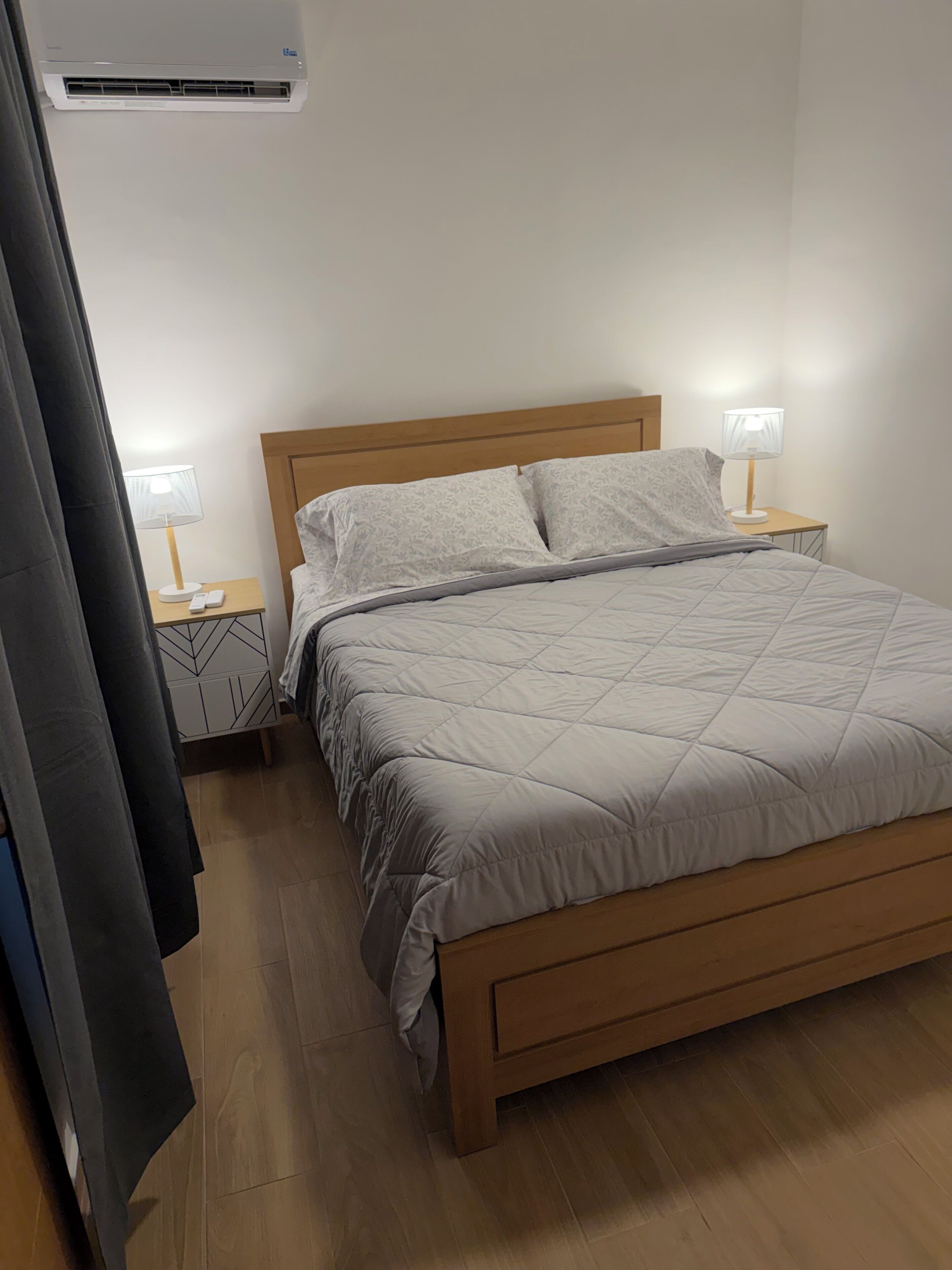 A neatly made bed with a wooden headboard, two bedside tables with lamps, and a wall-mounted air conditioner.