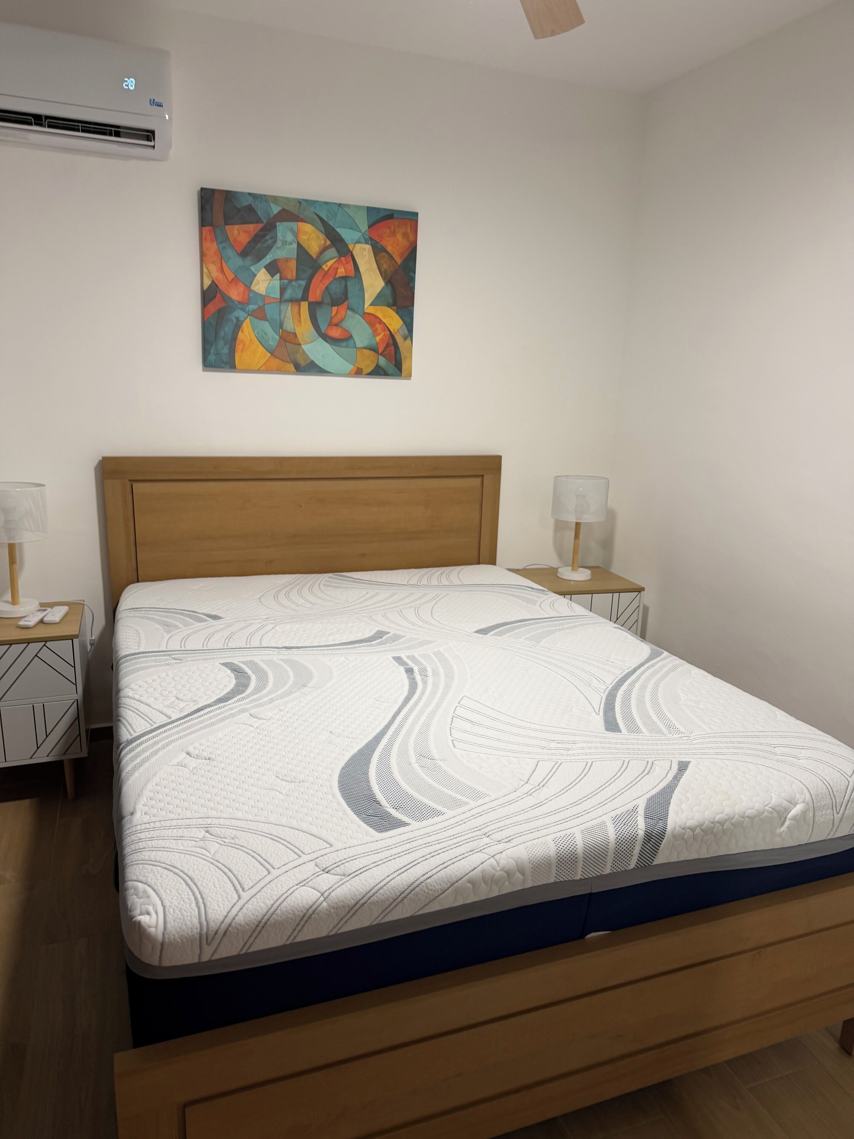 A bedroom with a wooden bed, a colorful abstract painting, and a white air conditioning unit.