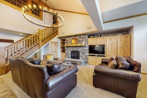 Ein geräumiges Wohnzimmer mit Kamin aus Stein, Ledersofas, einem Fernseher und einer Holztreppe.