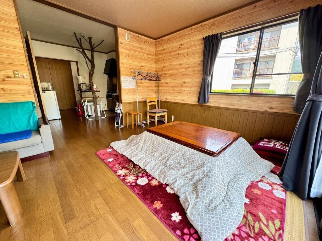 A wooden room with a bed, a table, and a chair. There is a window with curtains, a coat rack, and a small table with a chair.