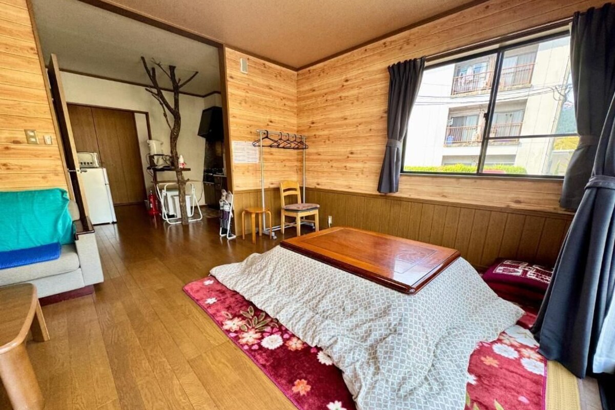A wooden room with a bed, a table, and a chair. There is a window with curtains, a coat rack, and a small table with a chair.
