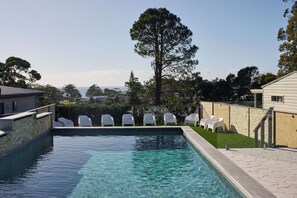 Une piscine à l’eau claire, entourée de chaises blanches, d’un grand arbre et d’un mur de pierre.