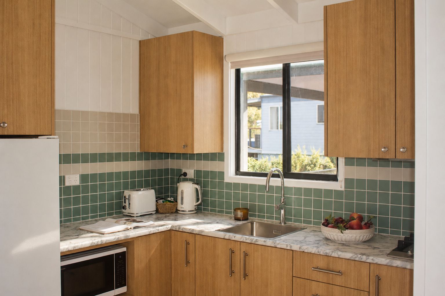 A kitchen with wooden cabinets, a white refrigerator, a sink, a window, and a countertop with a toaster, kettle, and a bowl of fruit.