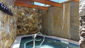 Un jacuzzi de paredes de piedra con una escalera metálica, techo de madera y piscina azul cristalina.