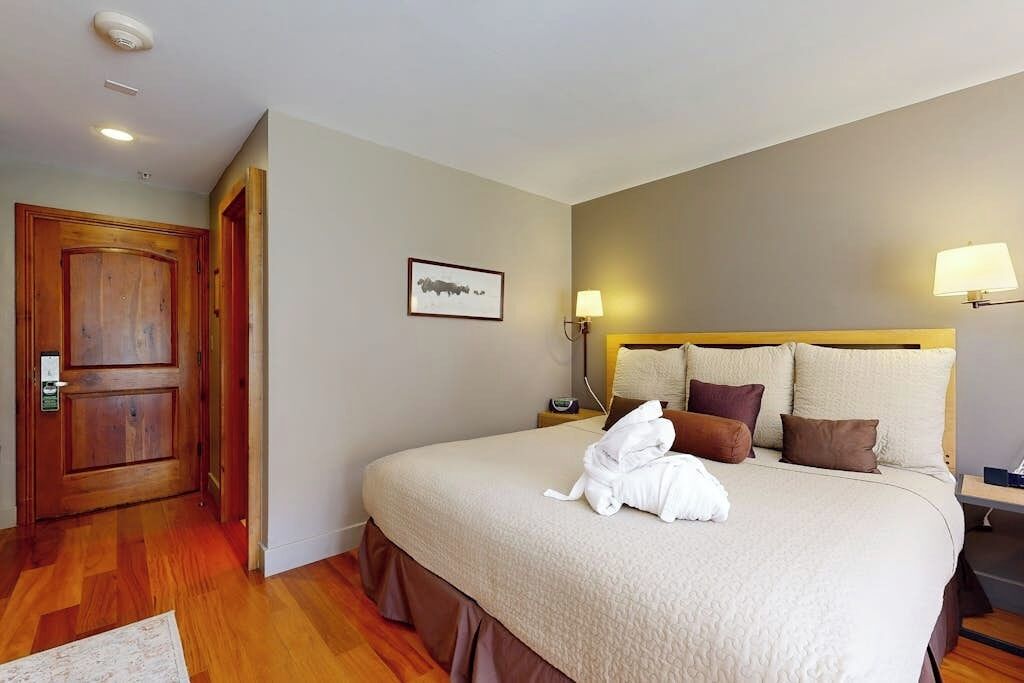 A hotel room with a bed, a wooden door, a framed picture on the wall, and a bedside lamp.